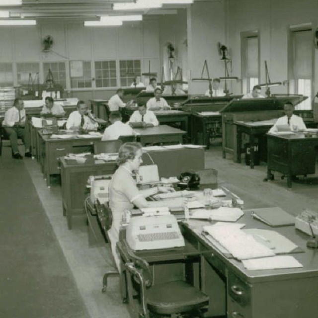 A black and white photo of an office space with employees working at their desks. 