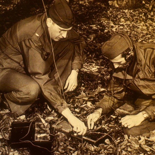 two men crouch down over radio in the woods; sepia photo
