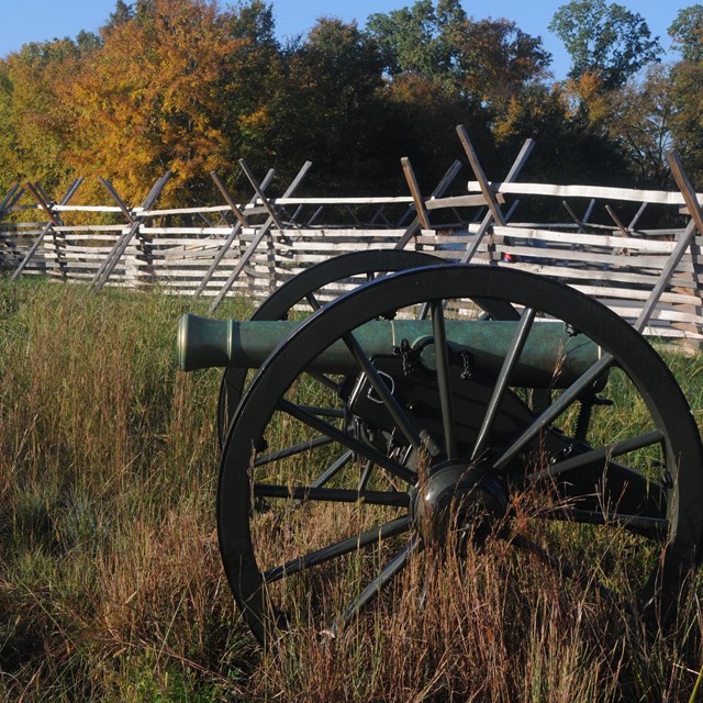 A weathered cannon stands in a field in front of a split rail fence and trees with fall color.