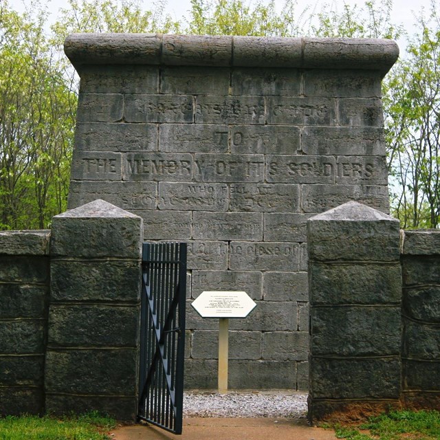 A grey stone wall with a iron gate stands in front of a cube-shaped stone structure.