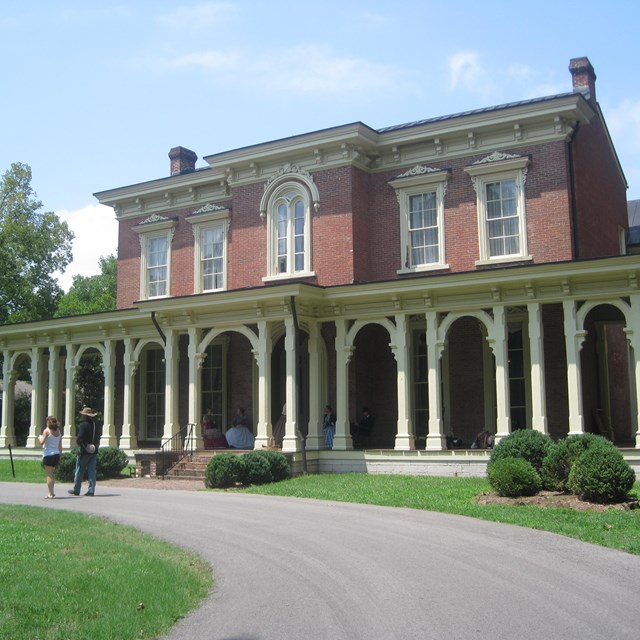 Two people walk in front of a Antebellum mansion made with bricks and thin wood columns.