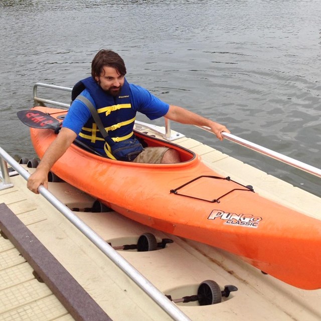 A man in a orange kayak uses an accessible kayak launch.