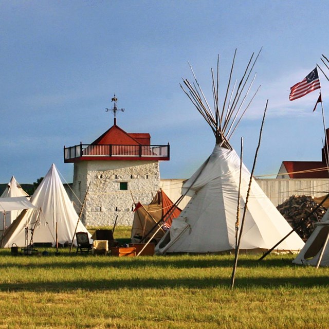 Green grass, blue cloudy sky, white tipis in front of a white fort wall and bastion with a red roof