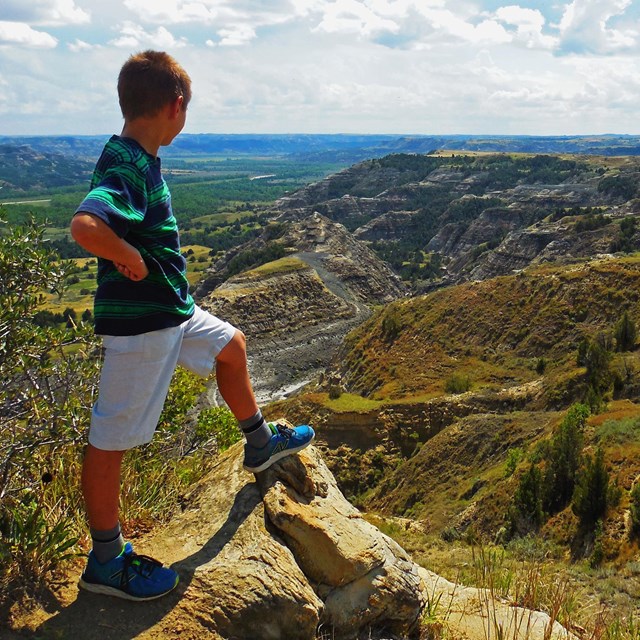 A child looks out into the sunny badlands with hands on hip and foot resting atop a rock. 