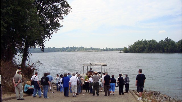 People stand on a boat ramp, waiting to board a small boat on a river.