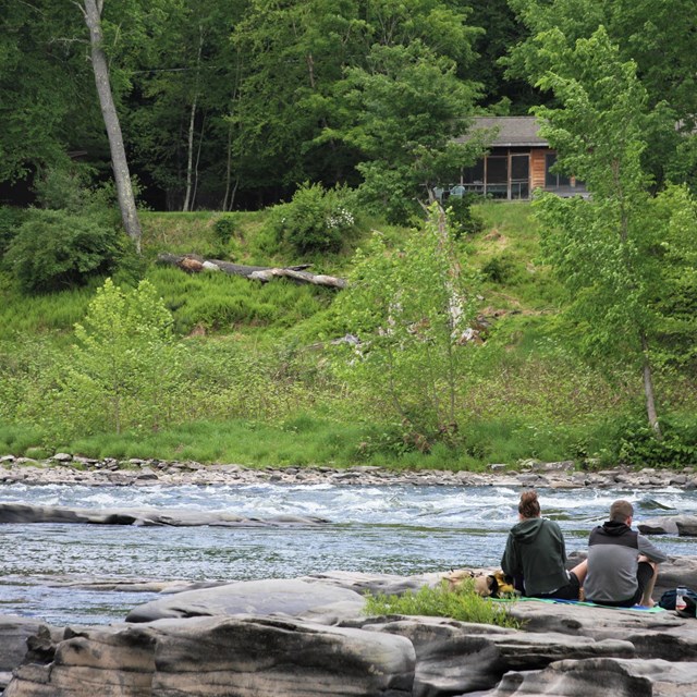 Couple sits on rocks on the edge of a river. Yellow-green trees form a forest across the river.