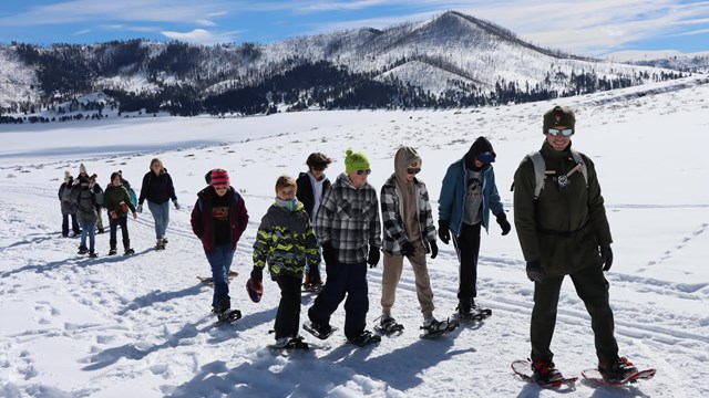 A park ranger leads a large group of students on a snowshoe hike.