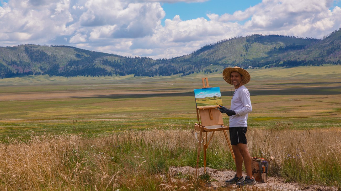 A painter stands next to an easel and a painted canvas at the edge of a vast grassland.