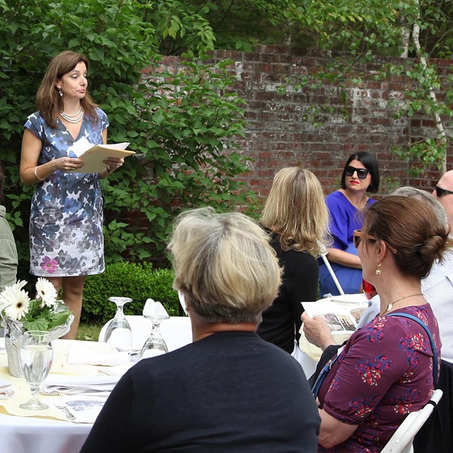 A woman speaks to a seated crowd in a garden.