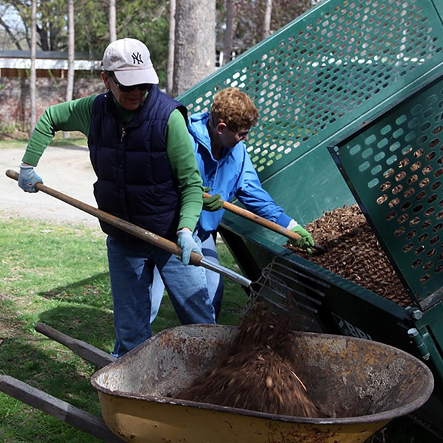 Two women shovel soil from a truck into a wheel barrow. 