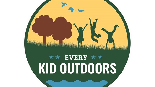 A round logo with kids jumping on green grass, brown trees and yellow sky.