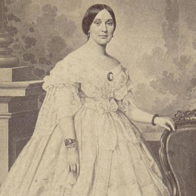 A picture of Varina Howell Davis standing in a white dress leaning on a chair. 