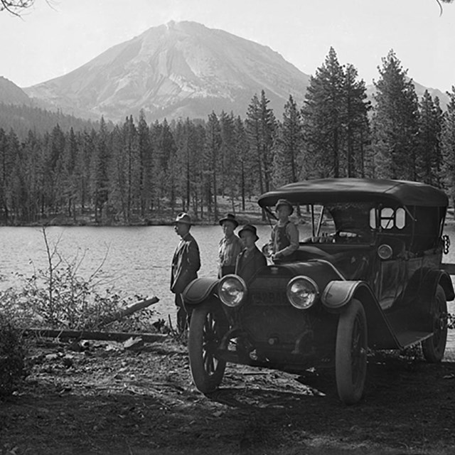 black and white photo of people standing by a car with lake and volcanic peak behind them