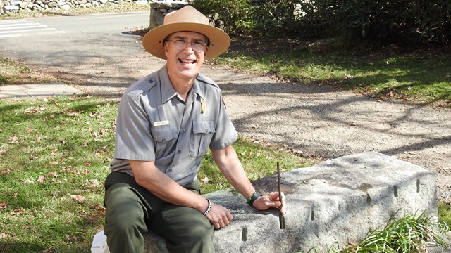 A park ranger sits on a low stone bench holding some small tools and smiling at the camera.
