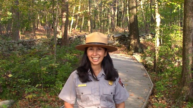 A park ranger smiles at the camera standing on a boardwalk in a green forest.