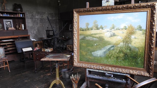 A large painting of a green landscape in a dark room filled with easels and artist tools.