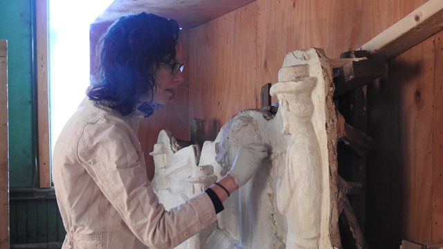 A person wearing gloves closely examines a large plaster figure.