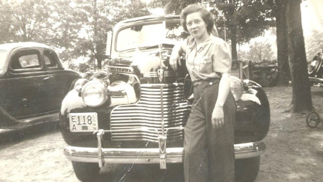 A black and white photo of a woman leaning up against a car.