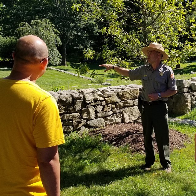 A park ranger stands in a group of people outside in a grassy field next to a stone wall.