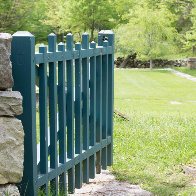 A green wood gate opens through a stone wall onto a grassy open field