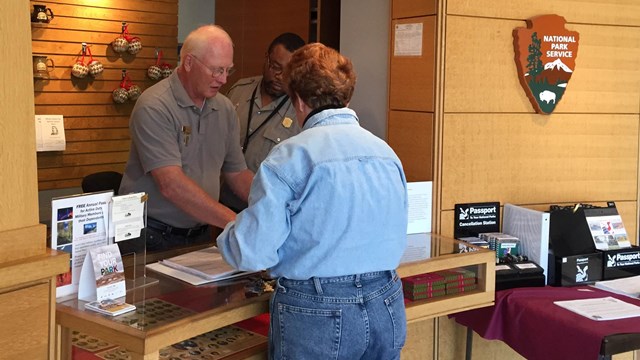 Two men behind a counter greet and talk to a woman in jeans and blue shirt