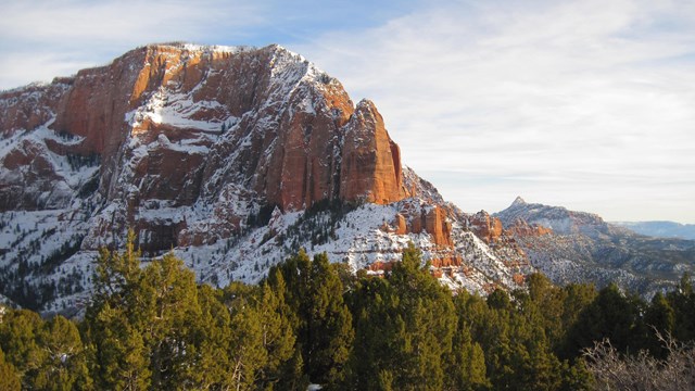 Red rock with snow on it extends high above a green forest.