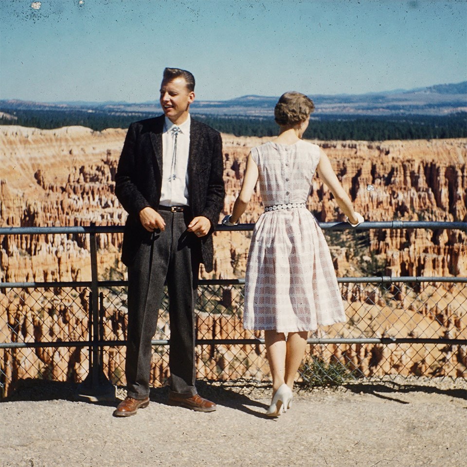 A couple in formal 1950s clothing stand at a railing overlooking a vast landscape of red rock spires and cliffs