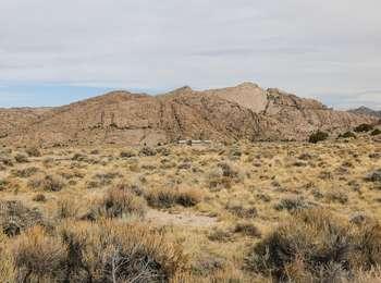 Behind a sagebrush and dried grass plain, rises a highly fractured, barren, tan and grey natural rock formation
