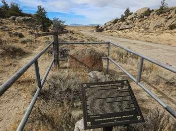 Grave with headstone, wooden cross, and a sign in a pass between two dry grass and sagebrush covered rocky ridges