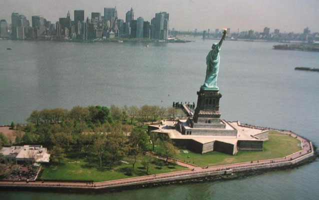 The Statue of Liberty stands on Liberty Island, with the skyline of New York in the distance.