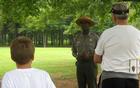A ranger and two visitors are standing under trees as the ranger begins his program.