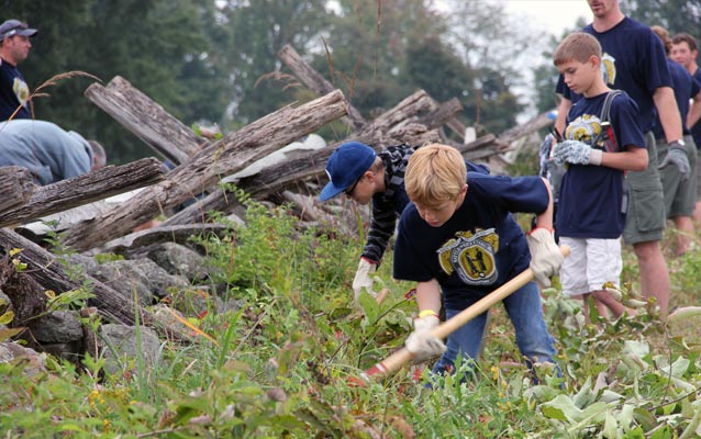 Young boy bends over to clear vegetation near a historic fence