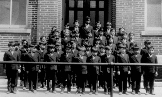 Children stand in suits in front of a schoolhouse