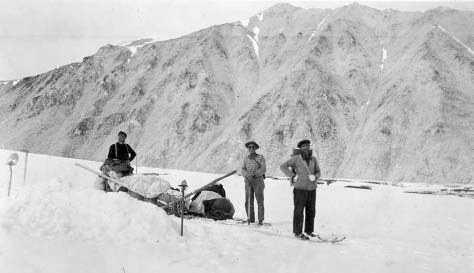 three men stnading on a snowy mountainside pulling a large sled