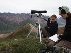 two women sitting on a mountaintop with a spotting scope