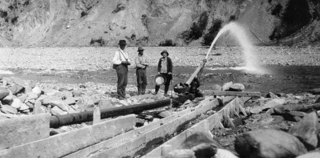 back and white image of three men standing near a hydraulic tube spewing water into a creek