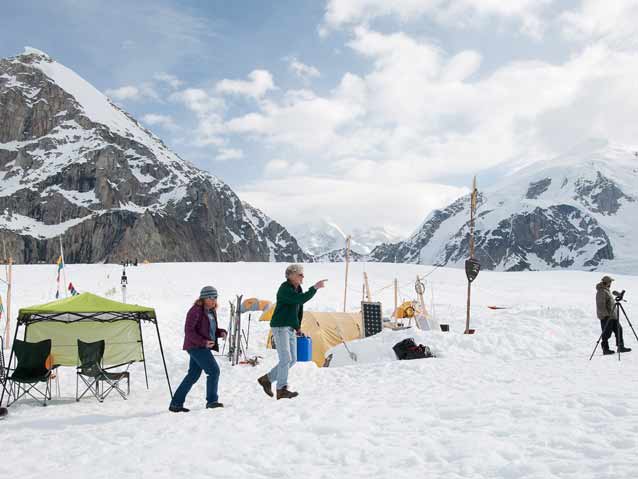 several people walking past tents set up on snow, surrounded by steep mountains