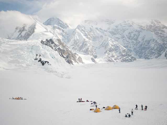 view from high above a cluster of tents in a vast snowy field, surrounded by mountains