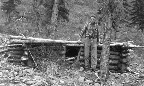 black and white image of a man standing near a very small log cabin 