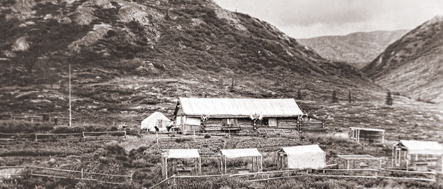 historic image of log cabin and small enclosures at the foot of a large hill