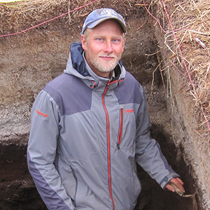 Researcher stands in archaeological dig with tools in hand