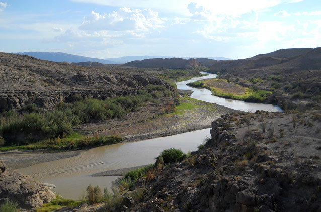 Brown water winds through an expansive valley below a partly cloudy sky.