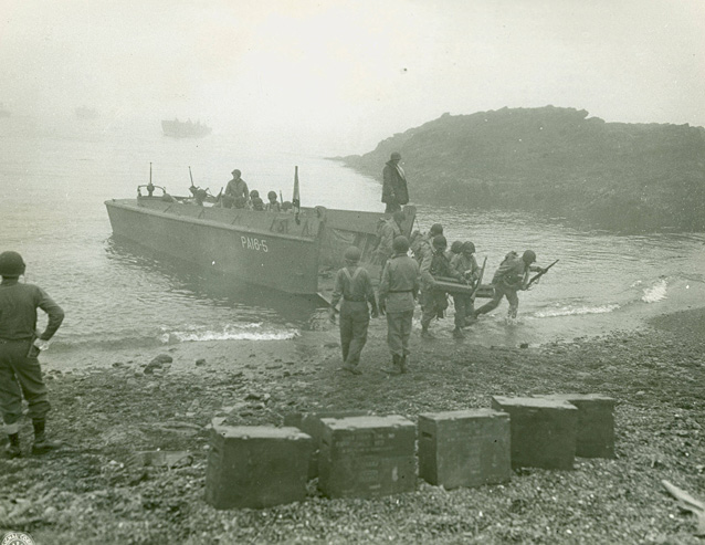 Uniformed soldiers carry gear off a boat onto a rocky beach.