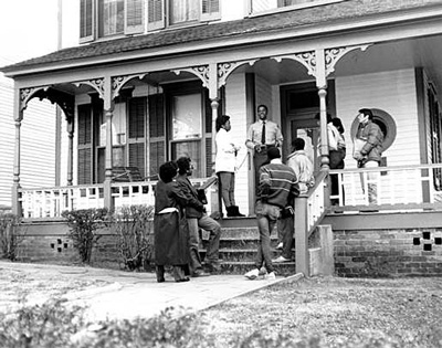 A tour guide stands on the porch of a house and speaks to a gathered group.