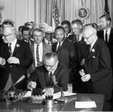 President Johnson, surrounded by group of people, signs a bill seated at desk