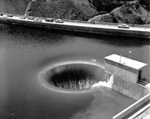 Black and white photo of a water pouring into a glory hole spillway