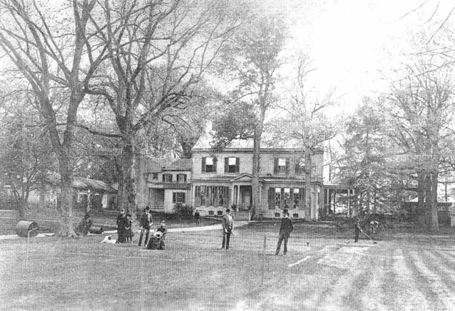 A group of people gather on a lawn in front of a large house in a black and white image. 