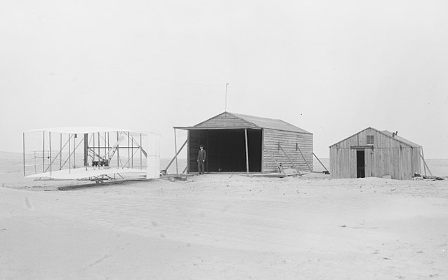 The Wright Flyer next to two camp buildings- Kitty Hawk, 1903