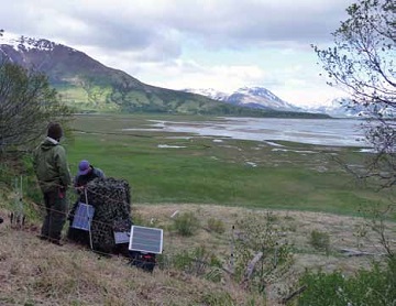 two people install a camera and solar panels with a floodplain and mountains in the distance