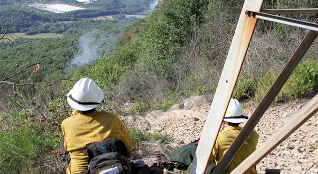 Two firefighters sit on a ridgetop overlooking a forested slope and smoke column.